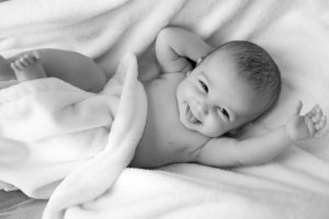 Smiling dark-haired baby boy lying supine on blanket with tongue out, left arm outstretched, and right hand behind head
