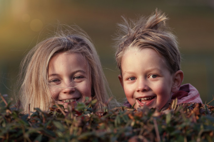 Two smiling blonde siblings, a girl and boy, have disheveled and windblown hair while peeking over a hedge