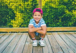 Young boy in a backwards red baseball cap and blue striped shirt crouches on a wooden bridge while smiling