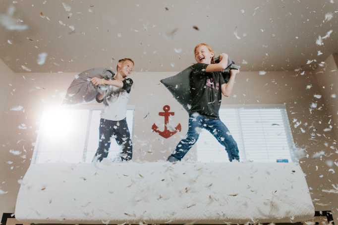 A young brother and sister smile and laugh as they have a pillow fight on a bed with feathers all around them in the air