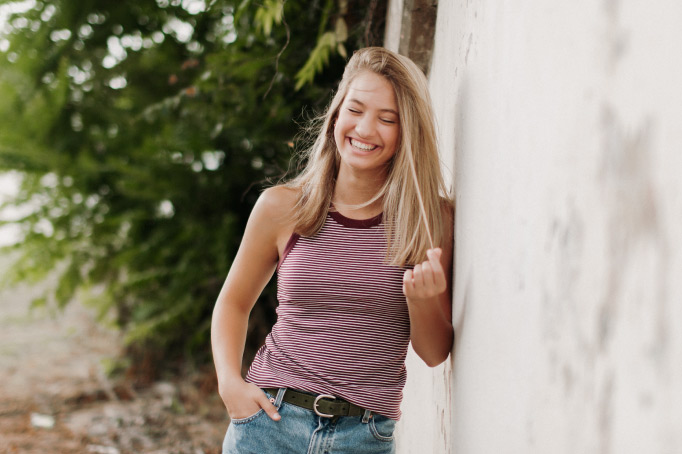 Blonde teenager who needs her wisdom teeth removed wears a pink tank-top while leaning against a white wall