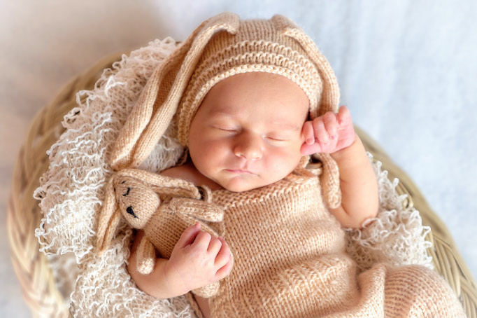 Aerial view of a sleeping baby wearing a crocheted sweater and rabbit ears beanie in a basket while holding a bunny