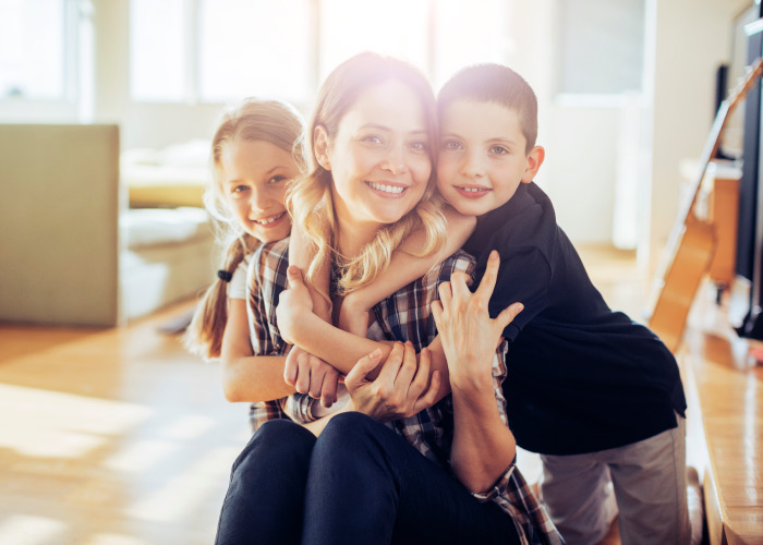 Mom smiles as she sits in a family room flooded with natural light, embraced by her young son and daughter