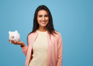 Dark-haired woman smiles and holds a piggy bank because she is going to use her dental insurance before the end of the year